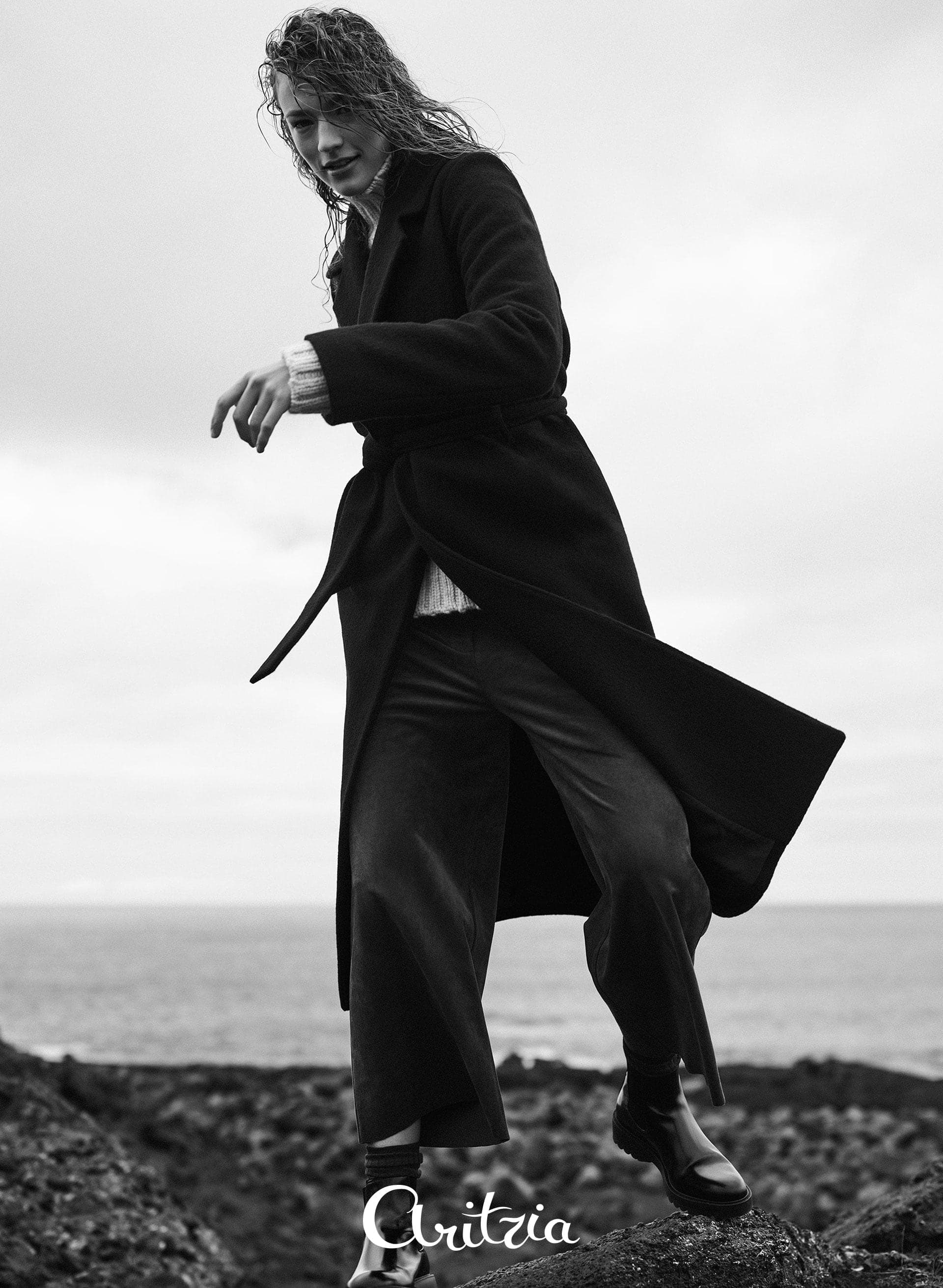 Black-and-white image of a model in a long coat on a rocky shore